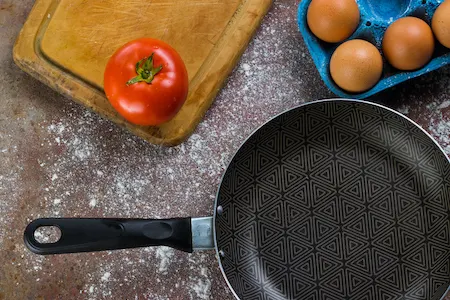 Frying pan, tomato, and carton of eggs on chopping block dusted with flour