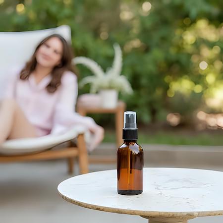 a person relaxing outside in the backyard on a lounge chair in the background and in the foreground a small amber glass spray bottle on a table