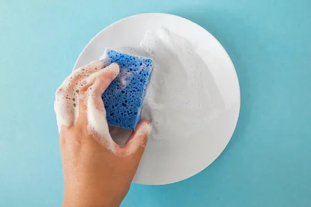 hand holds a plate with a cleaning sponge with soap foam on a blue isolation background