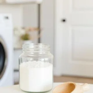 Picture of a clear glass jar filled with homemade laundry scent booster sitting on a table next to a small wooden scoop